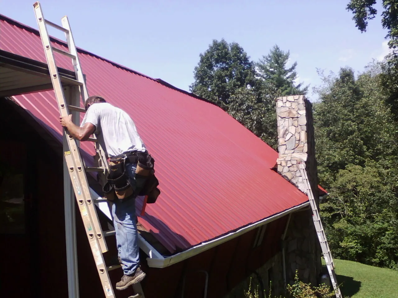 Roofer on a ladder beside a red metal roof and stone chimney — Soffit & Fascia Repair by Roofing Brothers in Promise City